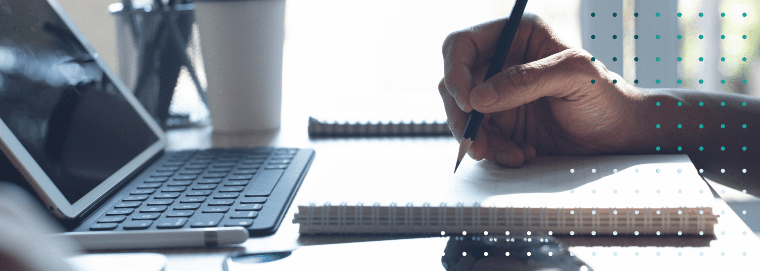 Image of a person's hand holding a pencil atop a piece of apper, in front of a computer screen.