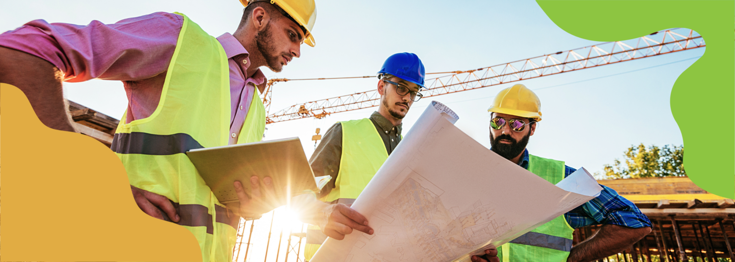 Three people stand looking at a large paper held by the personat the center. Each are wearing hard hats and reflective vests. There is building equipment behind them.