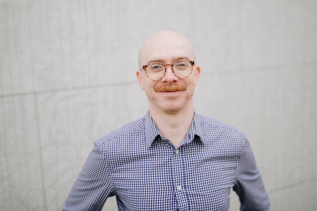 A bald man with glasses and a mustache, wearing a blue checkered shirt, stands in front of a light-colored textured wall, smiling at the camera.