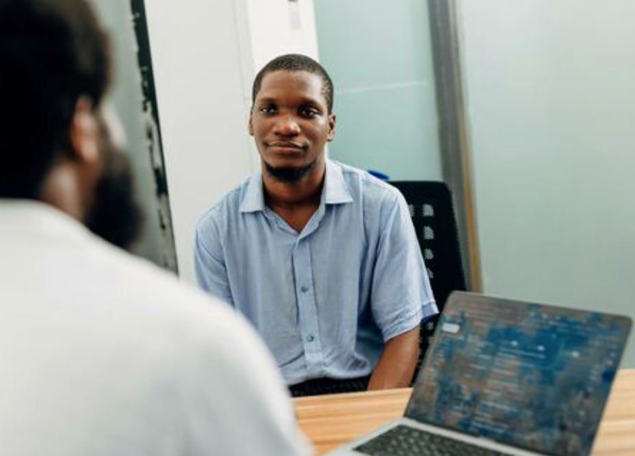 Two people sit across from each other at a table. One is wearing a blue collared shirt and the other has on a white shirt. There's a laptop on the table between them.