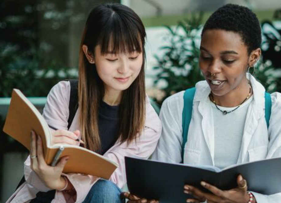 Two young adults sit side by side outdoors, leaning in over their open notebooks as they study together.