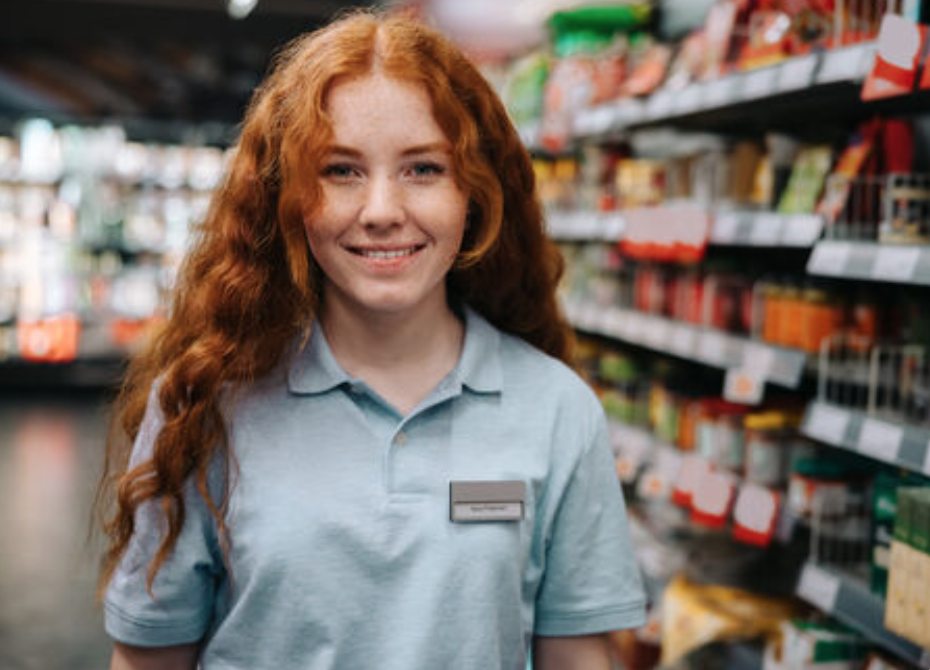 A red-haired girl in a blue collared shirt stands in front of a camera. She's wearing a name tag and is standing in a grocery store aisle.