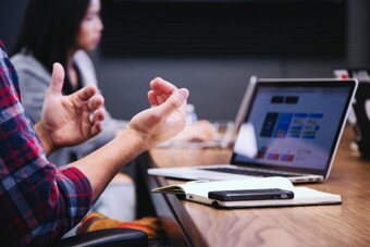 La imagen muestra un entorno profesional con dos personas en una mesa de conferencia. En primer plano, una persona está gesticulando con las manos mientras habla. Esta persona lleva una camisa de cuadros. Delante de ellos hay un cuaderno cerrado con un teléfono inteligente colocado encima. Una laptop está abierta sobre la mesa, mostrando una página web colorida o una interfaz de aplicación.
