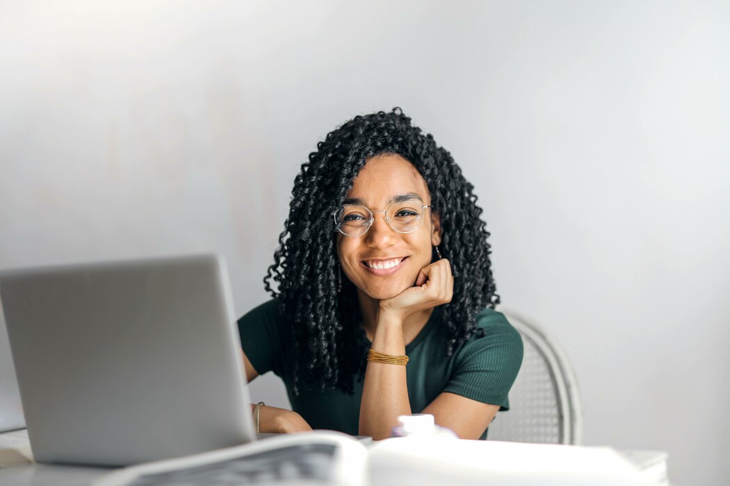 La imagen muestra a una persona con cabello rizado y gafas sentada en una mesa con una computadora portátil frente a ella. Llevan una camisa verde oscuro y están sonriendo, apoyando la barbilla en una mano. El fondo es de color blanco roto, lo que da a la escena una sensación brillante y aireada. 