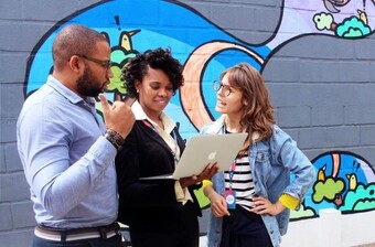 La imagen muestra a tres personas de pie frente a un mural colorido pintado en una pared de ladrillos. A la izquierda, una persona con una camisa azul claro gesticula con la mano mientras participa en una conversación. En el centro, una persona con cabello rizado, vestida con un blazer negro, sostiene una laptop abierta, pareciendo mostrar algo en la pantalla a los demás. A la derecha, una persona con gafas, una camisa a rayas y una chaqueta de mezclilla, mira atentamente la laptop con una mano en la cadera. 