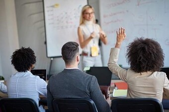 La imagen muestra un aula o sala de seminarios donde una persona está de pie al frente, frente a tres personas sentadas. La persona de adelante, con cabello claro, lleva gafas y una camiseta sin mangas. Parece que están hablando o presentando, con las manos haciendo gestos. Detrás de ellos, se ve una pizarra blanca con varios marcadores de colores y diagramas dibujados en ella. De los tres asistentes, dos han levantado la mano.