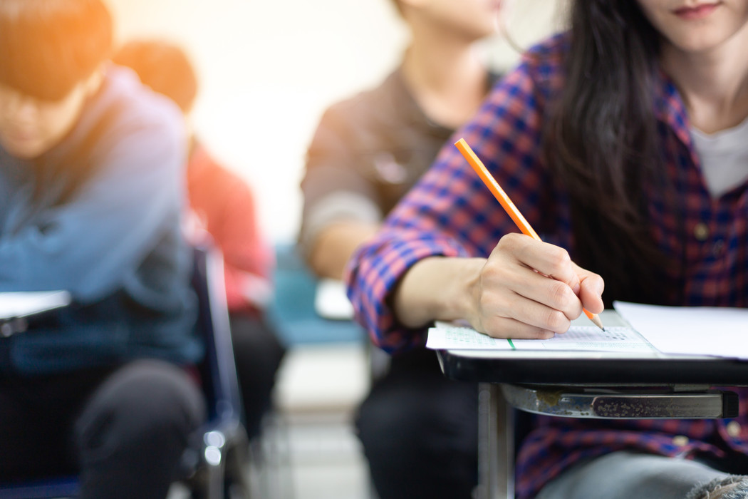 La imagen muestra un aula donde los estudiantes están sentados en escritorios tomando un examen. En primer plano, un estudiante con cabello largo y oscuro, que lleva una camisa de cuadros roja y azul, está concentrado en escribir con un lápiz en una hoja de examen colocada en un escritorio. El escritorio tiene un marco de metal negro con una superficie de escritura de madera