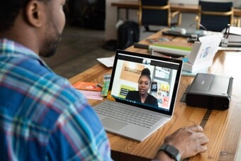 La imagen muestra a un hombre sentado en una mesa de madera, participando en una videollamada en una laptop. Él lleva una camisa de cuadros y un reloj en su muñeca izquierda. La laptop está sobre la mesa, mostrando a una mujer hablando en la ventana de chat de video. La mesa está llena de diversos objetos, incluyendo una impresora, una pila de papeles y algunos cuadernos coloridos.