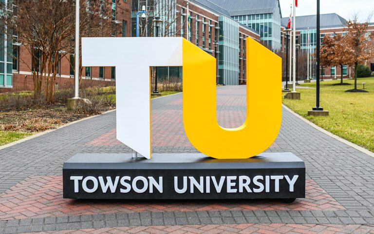 Una gran escultura independiente muestra las letras 'TU' — la 'T' en blanco y la 'U' en amarillo brillante — representando a Towson University. La escultura está sobre una base negra con 'TOWSON UNIVERSITY' escrito en blanco. Se encuentra en un camino peatonal pavimentado flanqueado por edificios de ladrillo, árboles y farolas en un campus universitario.