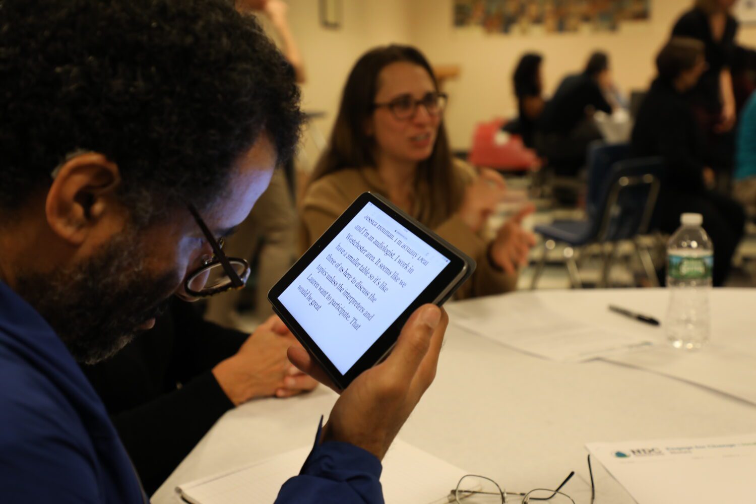 La imagen muestra a un hombre enfocado en el lado izquierdo, sosteniendo y leyendo de una tableta. La pantalla de la tableta muestra texto en una fuente clara y legible. El hombre tiene el cabello rizado corto, gafas y lleva una chaqueta azul. Él está sentado en una mesa cubierta con un mantel blanco.
