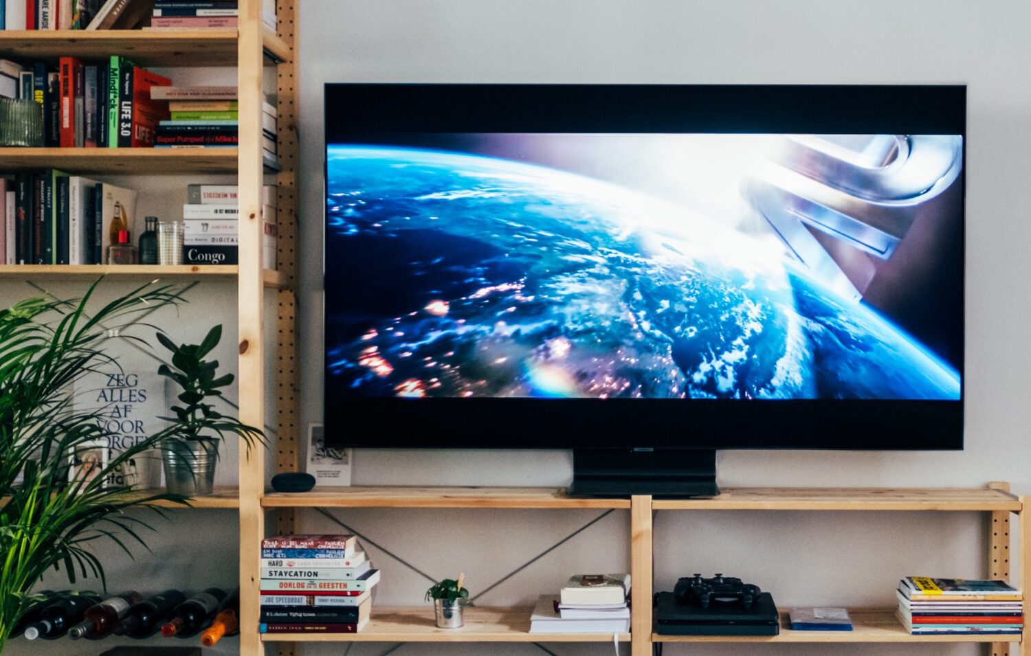 La imagen muestra una sala de estar con un televisor de pantalla plana grande montado en una estantería de madera. La televisión muestra una imagen de la Tierra vista desde el espacio, con la luz del sol iluminando una parte del planeta. En las estanterías que rodean la televisión, hay numerosos libros dispuestos tanto vertical como horizontalmente, y algunos objetos personales como una planta pequeña y controles de videojuegos.