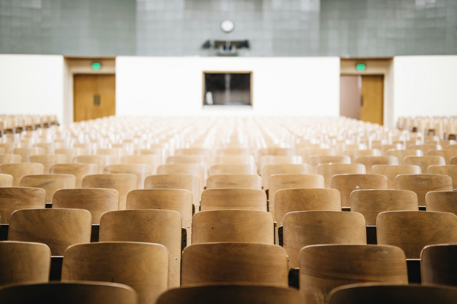 Rows of empty wooden seats in a large lecture hall with a clock at front.
