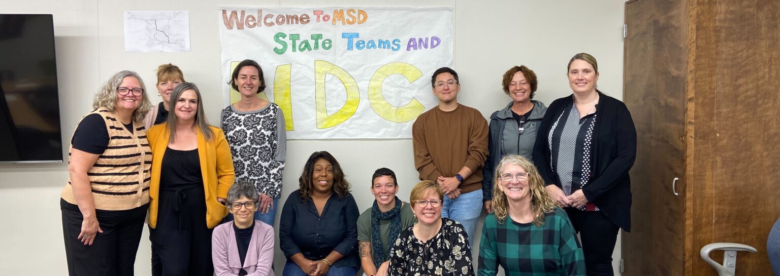The image shows a group of eleven people posing indoors against a wall. They are smiling and appear to be part of a professional or semi-formal gathering. In the background, a sign reading "Welcome to MSD State Teams and NDC" is visible, written in colorful letters. The group includes both men and women of various ages, with some seated in front and others standing behind them. A television is mounted on the left side of the room, and a wooden piece of furniture is on the right. The walls are light-colored, contributing to a neutral backdrop.