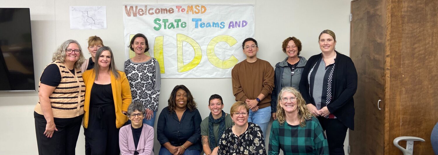 The image shows a group of eleven people posing indoors against a wall. They are smiling and appear to be part of a professional or semi-formal gathering. In the background, a sign reading "Welcome to MSD State Teams and NDC" is visible, written in colorful letters. The group includes both men and women of various ages, with some seated in front and others standing behind them. A television is mounted on the left side of the room, and a wooden piece of furniture is on the right. The walls are light-colored, contributing to a neutral backdrop.