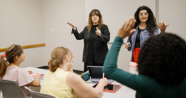 An instructor with an interpreter signing in a class while students sit at a table, one raising a hand.