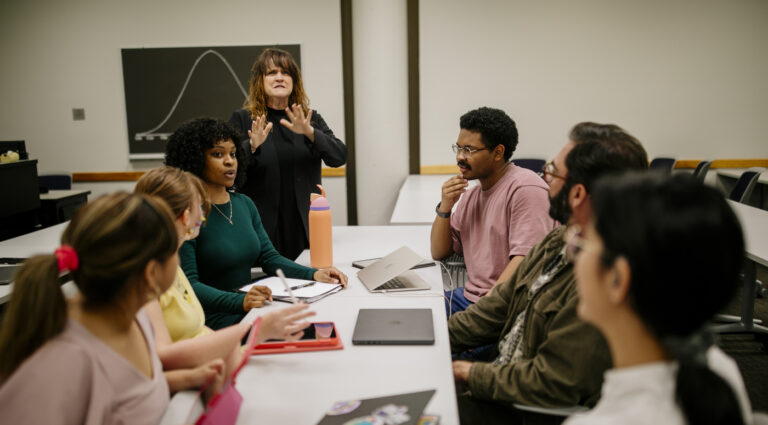 Group of students at a classroom table with laptops while an instructor uses American Sign Language near a bell-curve chalkboard.