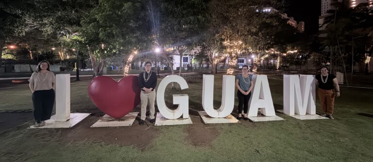 Four people pose at night beside a large 'I heart GUAM' sign in a park with trees and string lights.