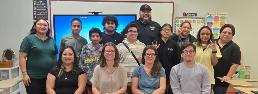 Group of students and teachers in a classroom, some signing I love you in ASL, with an ASL alphabet banner above a smartboard.