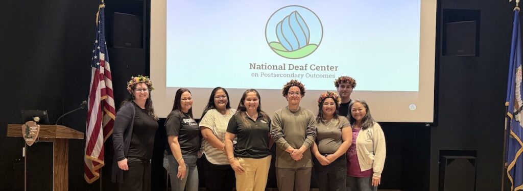 Group of nine people posing in front of a National Deaf Center presentation screen, with flags and podium on stage.