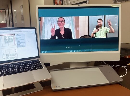 Desk setup with monitor showing two people (Molly and Kelton) on a video call flashing peace signs, beside an open laptop, keyboard, and mouse.