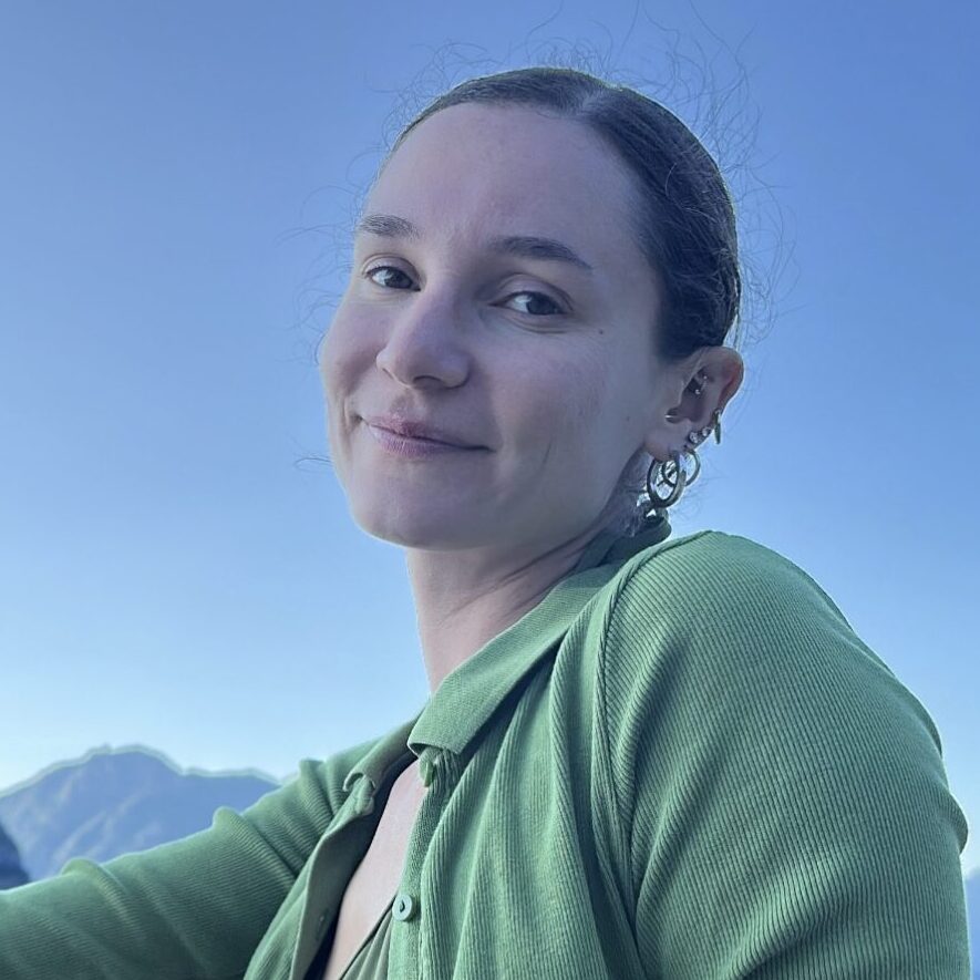 Smiling person in a green cardigan outdoors with mountains beneath a clear blue sky.