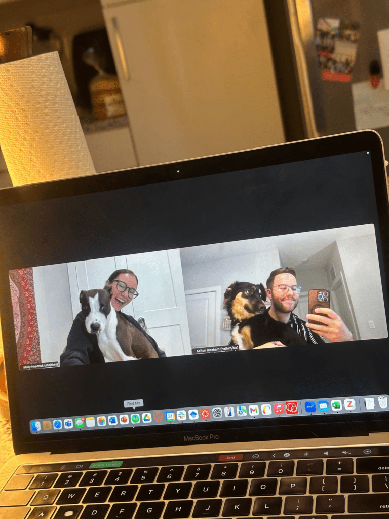 Laptop screen shows a Zoom call with two people smiling and holding dogs, in a cozy kitchen setting.