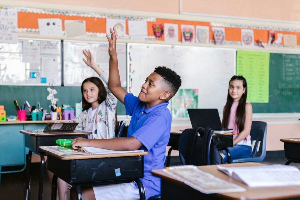 Elementary students seated at desks in a bright classroom, with two raising their hands to answer a question.