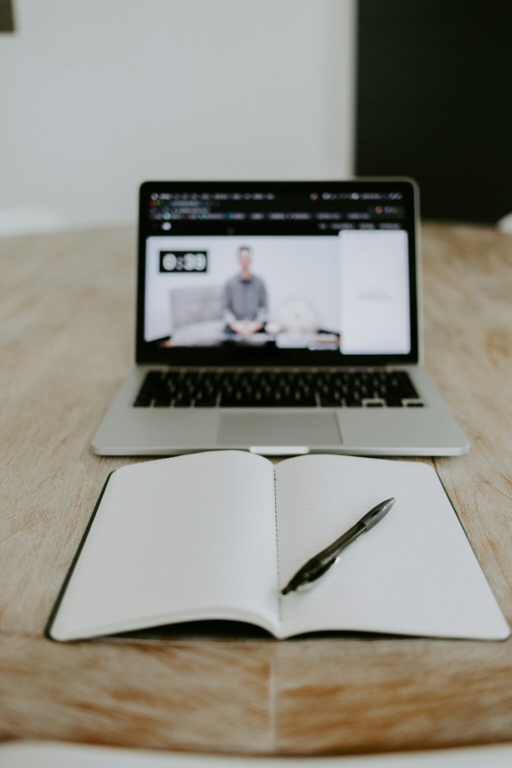 Open notebook and pen on a wooden table in front of a laptop streaming a blurred video lesson.