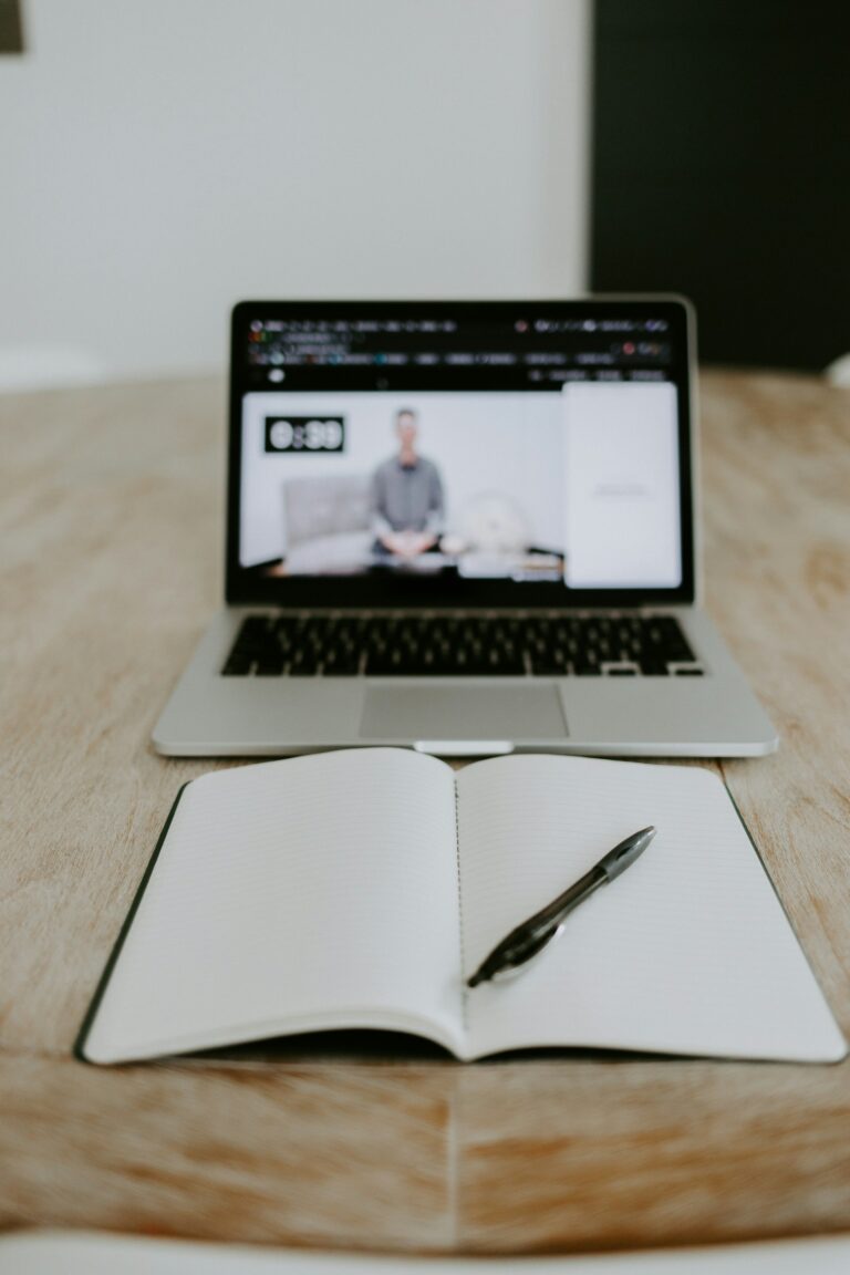 Open notebook and pen on a wooden table in front of a laptop streaming a blurred video lesson.