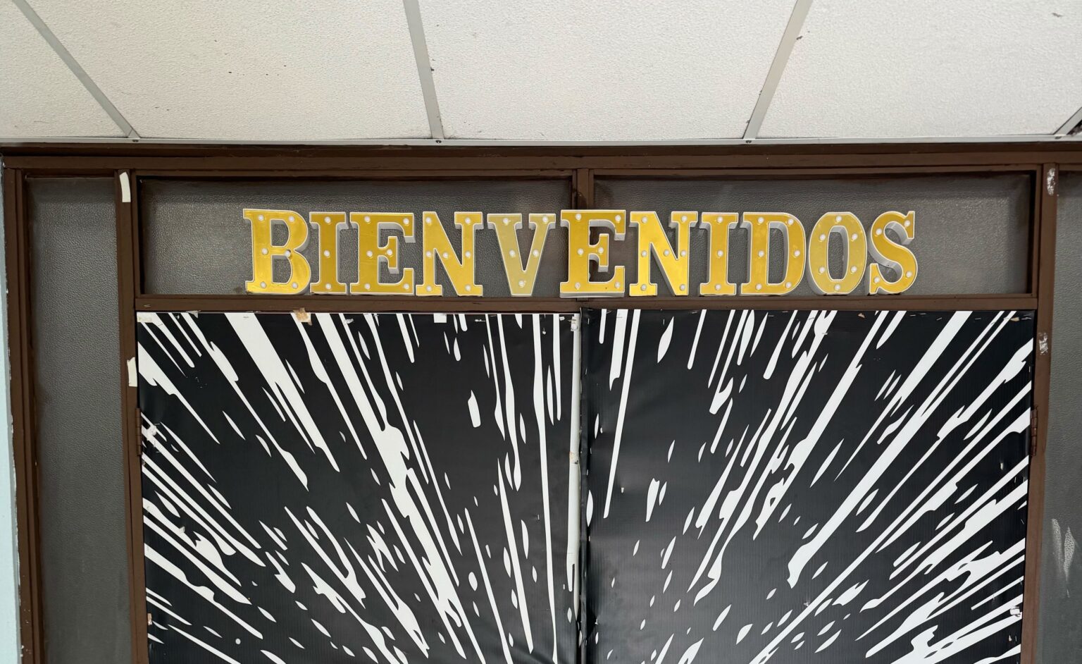 A pair of double doors wrapped in a bold black-and-white starburst design, topped with gold marquee-style letters spelling "BIENVENIDOS.”