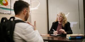 A student uses American Sign Language during a one-on-one academic support session with a tutor seated across the table. The tutor holds a pen and takes notes as they communicate directly.