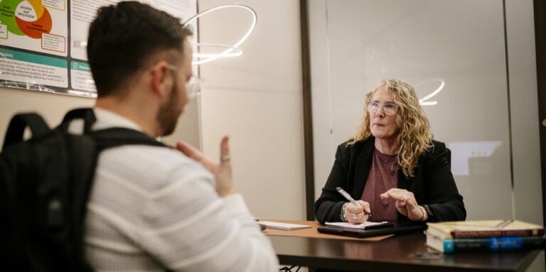 A student uses American Sign Language during a one-on-one academic support session with a tutor seated across the table. The tutor holds a pen and takes notes as they communicate directly.