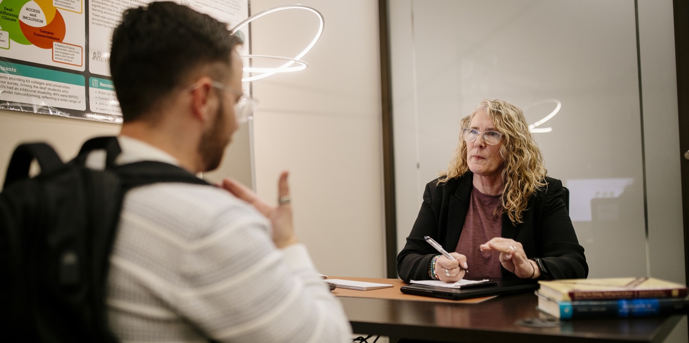 A student uses American Sign Language during a one-on-one academic support session with a tutor seated across the table. The tutor holds a pen and takes notes as they communicate directly.