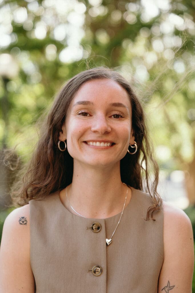 Smiling person with long hair wearing a beige top and heart necklace.