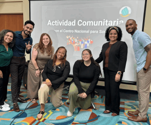Kate, Tia, and four other people from the community event in Aguadilla are smiling toward the camera in front of a screen that reads "Actividad Comunitario.