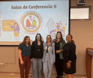 The image shows five people standing in front of a projector screen in a conference room. The screen displays a colorful presentation with the text "Salas de Conferencia II" at the top. The central graphic is a logo for "DivED" of "UPR Aguadilla," featuring an open book with wireless signal waves above it and the year 2002. Surrounding the logo are illustrations, including a person pointing at a board and another working at a desk.