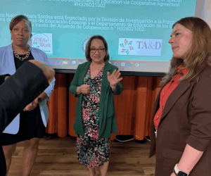 The image shows three people standing in front of a projected screen displaying informational text. The person in the center is wearing a green dress with a floral pattern and a green jacket. They are gesturing with their hand. To the left, a person in a light blue blouse and black skirt stands with their arms crossed. To the right, another person is wearing a brown jacket over a red shirt with beige pants. The background screen displays text and logos related to the National Deaf Center.