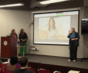 Tia and Kate showing a video during presentation with an interpreter standing on the other side of the screen. There is a group of people in the audience.