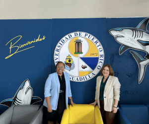 Tia and Kate smiling, standing in front of the seal of UPR Aguadilla on a mural.